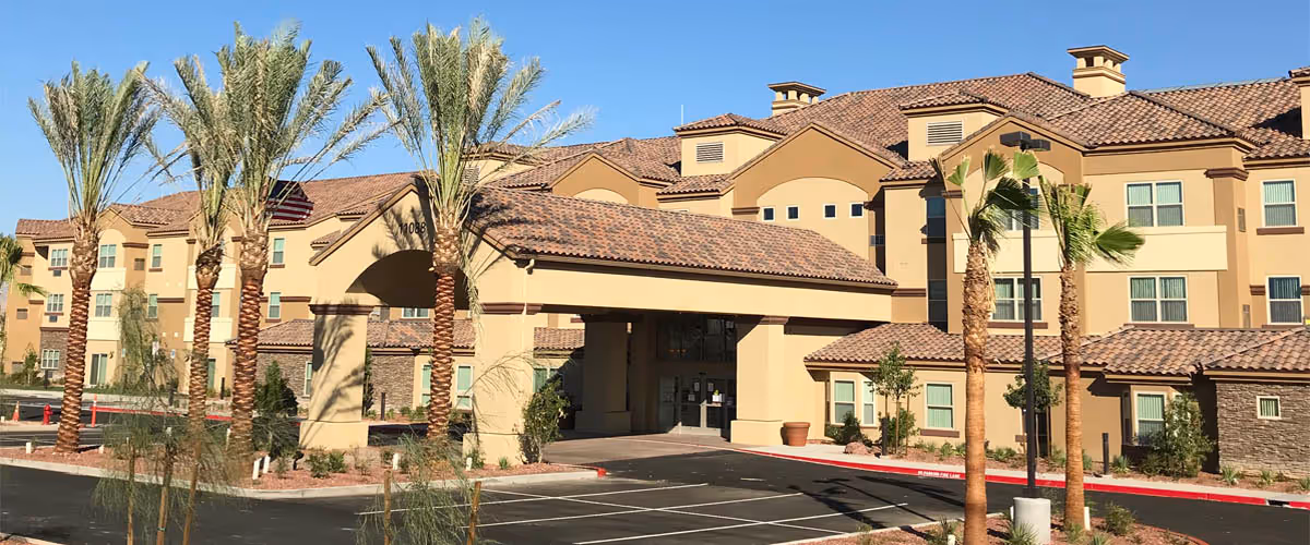 Exterior view of a large, multi-story retirement resort building with a covered entrance, palm trees, and a parking lot in front under a clear blue sky.