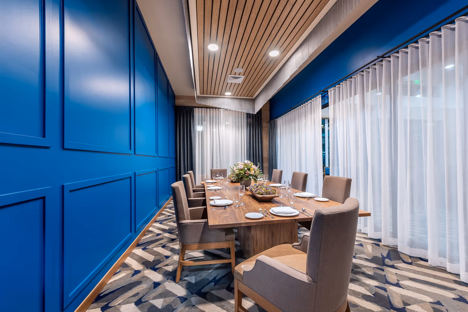 A modern dining room with a long wooden table set for eight people. The chairs are upholstered in beige fabric. The room features a blue paneled wall on one side and large windows with sheer white curtains on the other. The ceiling has wooden slats with recessed lighting, and the floor is covered with a patterned carpet.