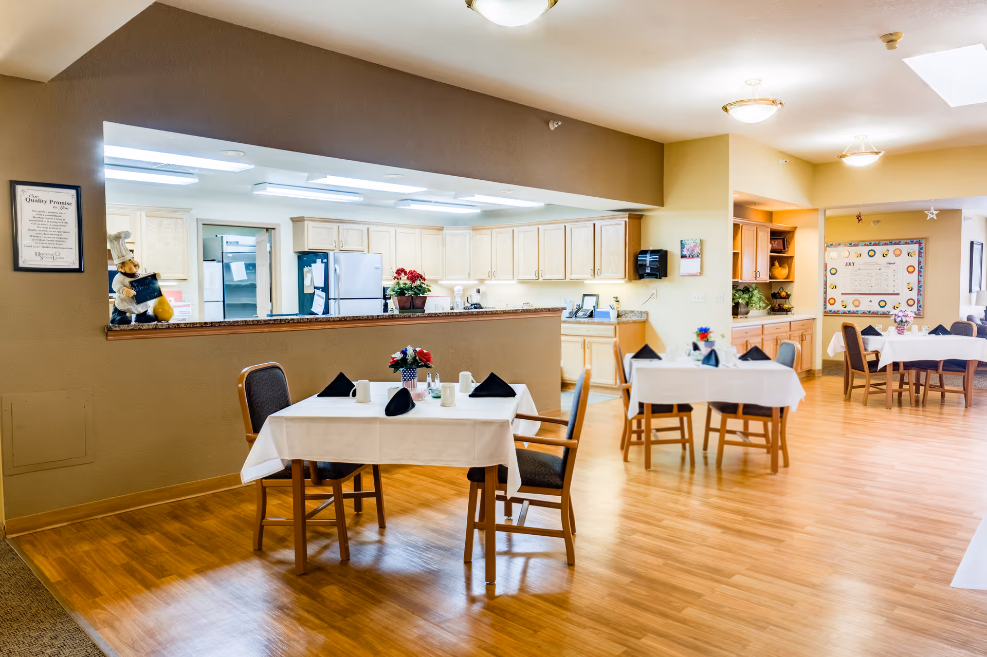A bright dining area in an assisted living facility with wooden floors and several tables covered with white tablecloths, each set with black napkins, cups, and small flower arrangements. The background shows a kitchen area with light wood cabinets, a refrigerator, and a counter with decorative items. The walls are painted in warm tones, and there is a bulletin board with a calendar on the far wall.