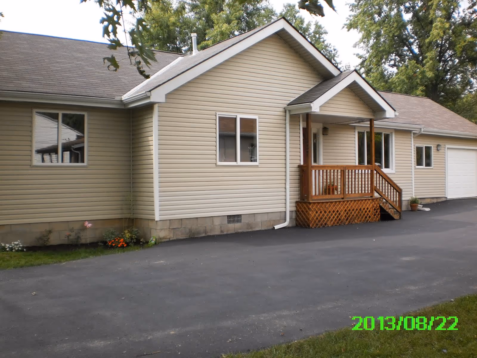 Exterior view of a single-story beige house with a small wooden porch and steps leading to the front door, surrounded by trees and a paved driveway.