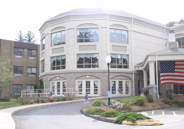Front exterior of a rehabilitation care building with a curved multi-story facade, large windows, an entrance ramp, American flags, and a circular driveway.