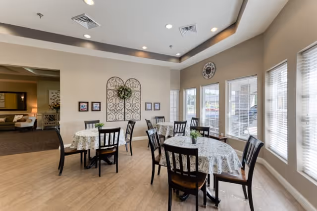 A bright dining area with round tables covered in floral tablecloths and surrounded by black wooden chairs. Large windows with blinds allow natural light to fill the room. The walls are beige with decorative wall art and a clock above the windows. The floor is light wood, and there is an adjacent lounge area visible in the background.