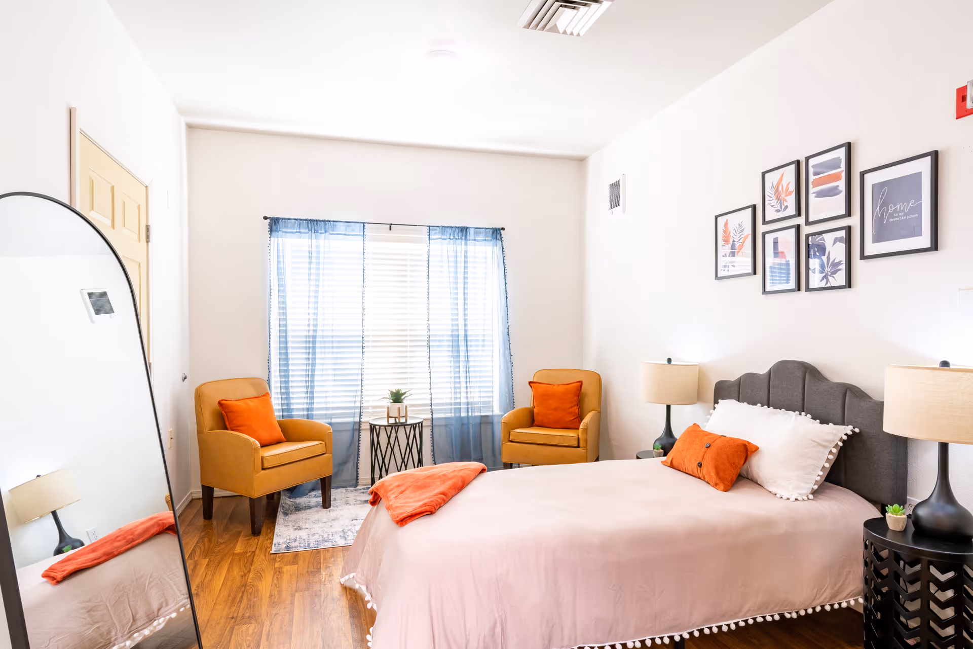 A bright furnished bedroom featuring a bed with orange pillows, two orange-accented armchairs by a window, bedside lamps and framed wall art.