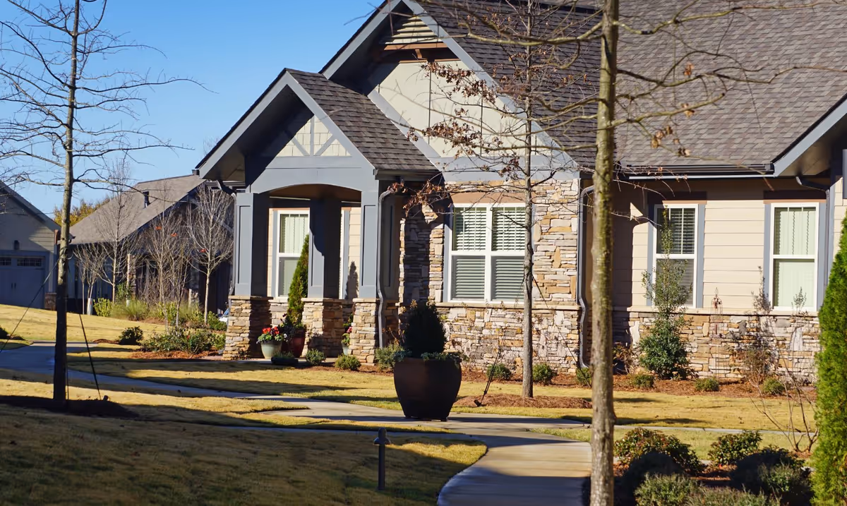 Exterior view of a residential building with stone and siding facade, a covered porch, and several windows. The building is surrounded by a landscaped yard with grass, small trees, shrubs, and a curved concrete walkway leading to the entrance.