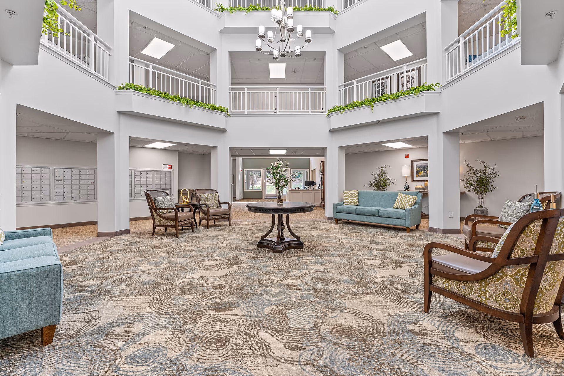 Spacious and well-lit interior common area of a senior living facility with a high ceiling and two levels of balconies. The room features patterned carpet flooring, a round wooden table with a flower arrangement in the center, several upholstered chairs and sofas arranged around the space, and mailboxes along the left wall. Green plants are placed on ledges and in vases, adding a touch of nature to the modern, clean design.
