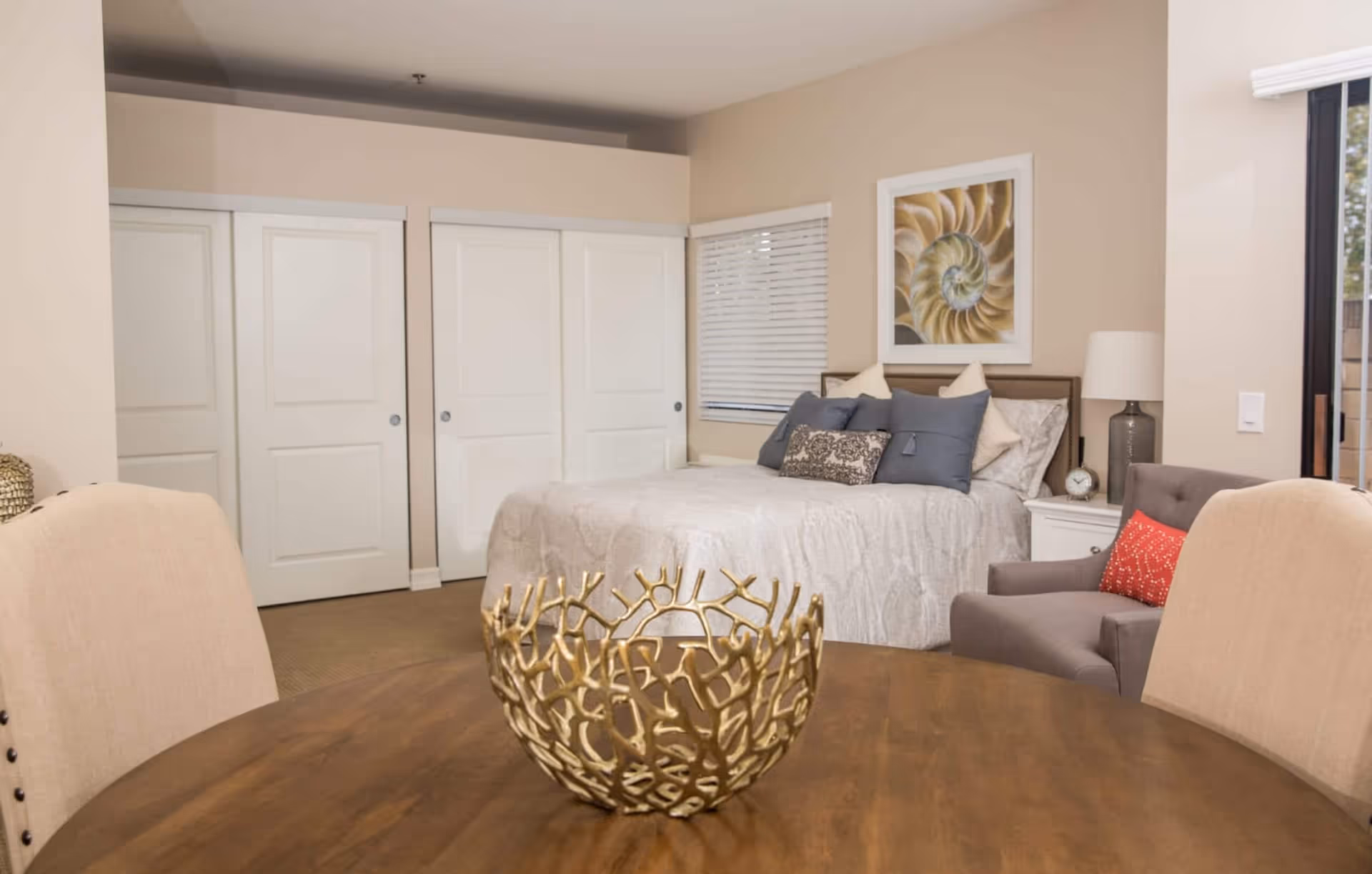 A bright studio-style bedroom with a made bed, bedside table and lamp, armchair with pillow, round dining table with a decorative gold bowl, and sliding closet doors.