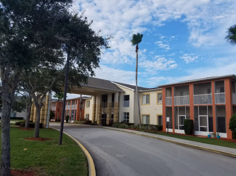 Exterior view of a senior living facility named Century Oaks with a curved driveway, green lawns, several trees including palm trees, and a two-story building with balconies and screened porches under a partly cloudy sky.