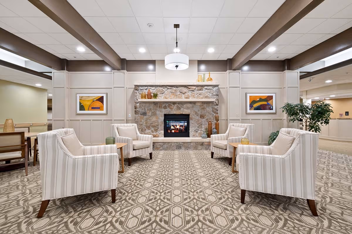 A cozy sitting area in a senior living facility featuring four striped armchairs arranged around a stone fireplace with a lit fire. The room has patterned carpet, beige paneled walls with colorful framed artwork, a hanging ceiling light, and decorative vases on the fireplace mantel. There is a potted plant to the right and a dining area visible to the left.