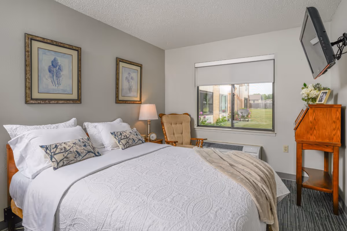 A neatly made bed with white bedding and decorative pillows in a bedroom. There are two framed floral pictures on the wall above the bed, a wooden rocking chair with a cushion, a bedside table with a lamp and clock, a window with a view of a grassy outdoor area, and a wooden desk with flowers and a photo frame. A flat-screen TV is mounted on the wall.