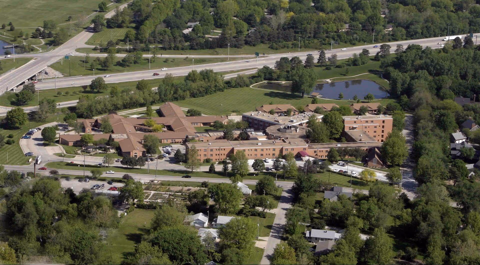 Aerial view of Topeka Presbyterian Manor, a large senior living facility surrounded by trees, roads, and residential houses. The building complex includes multiple connected structures with brown roofs and a small pond nearby.
