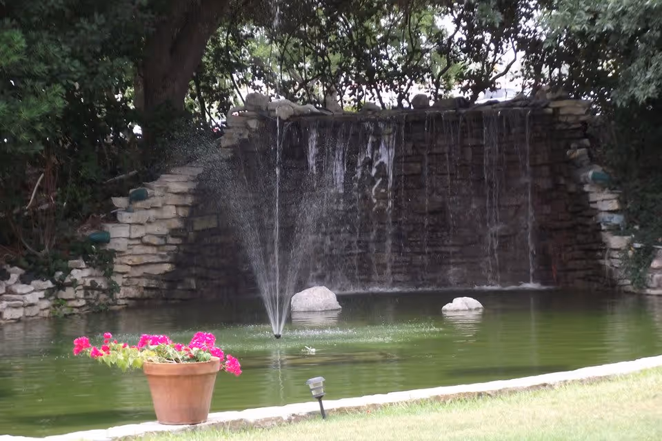 A landscaped pond with a central spray fountain and stone waterfall, with a potted pink flower in the foreground.