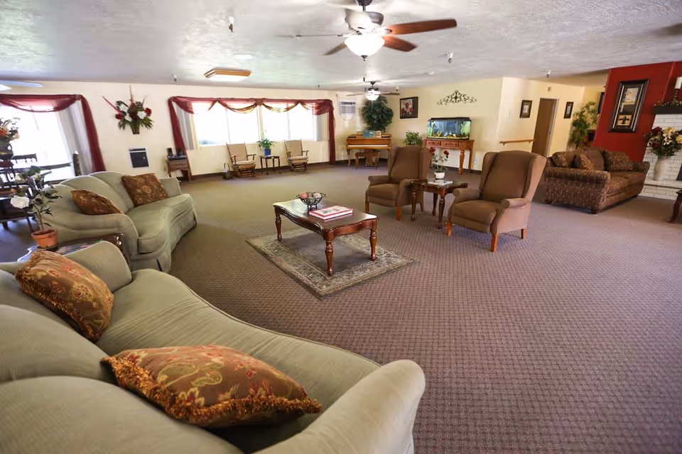 A spacious living room area in an assisted living facility featuring multiple upholstered sofas and armchairs arranged around a wooden coffee table with books and decorative items. The room has large windows with sheer curtains, ceiling fans with lights, a piano, an aquarium on a wooden stand, and various plants and floral decorations. The carpet is patterned, and the walls are painted in warm tones with framed artwork and a fireplace visible.