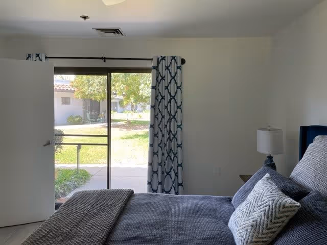 A bedroom with a neatly made bed featuring gray and white pillows and a gray blanket. There is a nightstand with a lamp next to the bed. A sliding glass door with patterned curtains opens to a green outdoor area with trees and a building visible in the background.