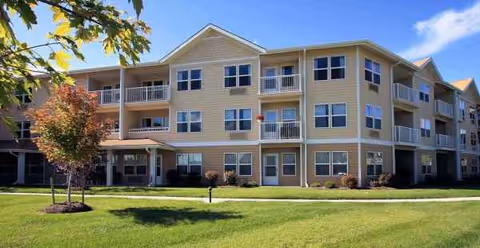Three-story beige senior living building with balconies and a grassy front lawn.