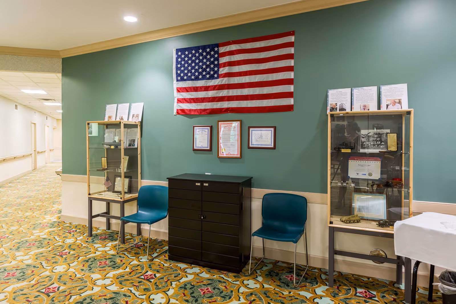 Interior hallway of a senior living facility with a green wall displaying an American flag and framed certificates. Two glass display cases with memorabilia flank a black cabinet, and two blue chairs are placed on either side of the cabinet. The floor has a patterned carpet and the hallway extends to the left.