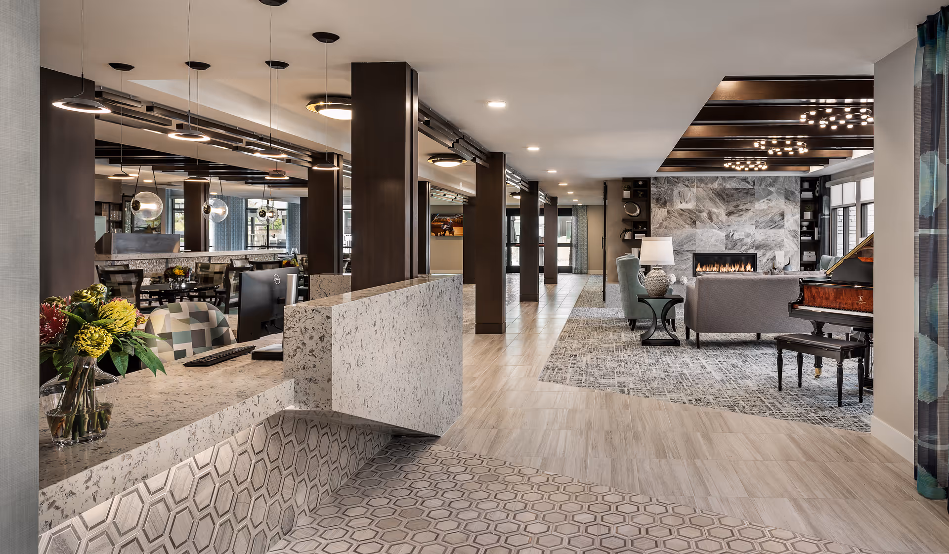Modern senior living facility interior with a reception desk on the left featuring a vase of flowers, a seating area with a fireplace and a grand piano on the right, and a dining area visible in the background. The space is well-lit with ceiling lights and has a mix of tile and carpet flooring.