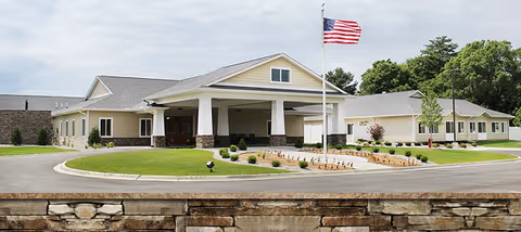 Front exterior of a senior living facility with a covered entrance, circular driveway, landscaped grounds, and an American flag on a flagpole.