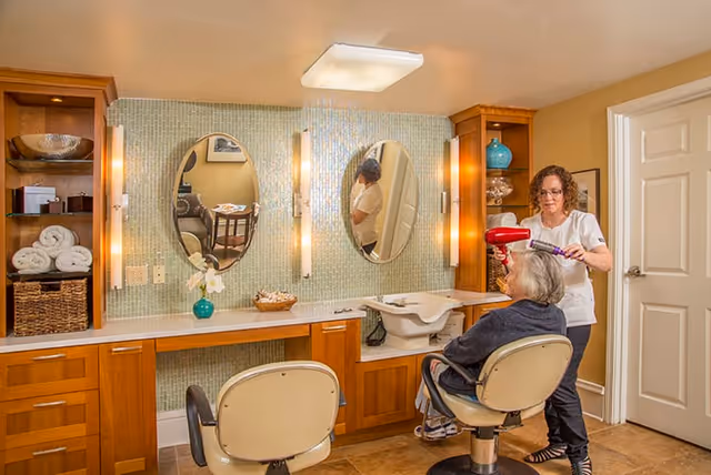 A caregiver blow-dries an elderly woman's hair in a salon area with oval mirrors, wood cabinetry, and salon chairs.