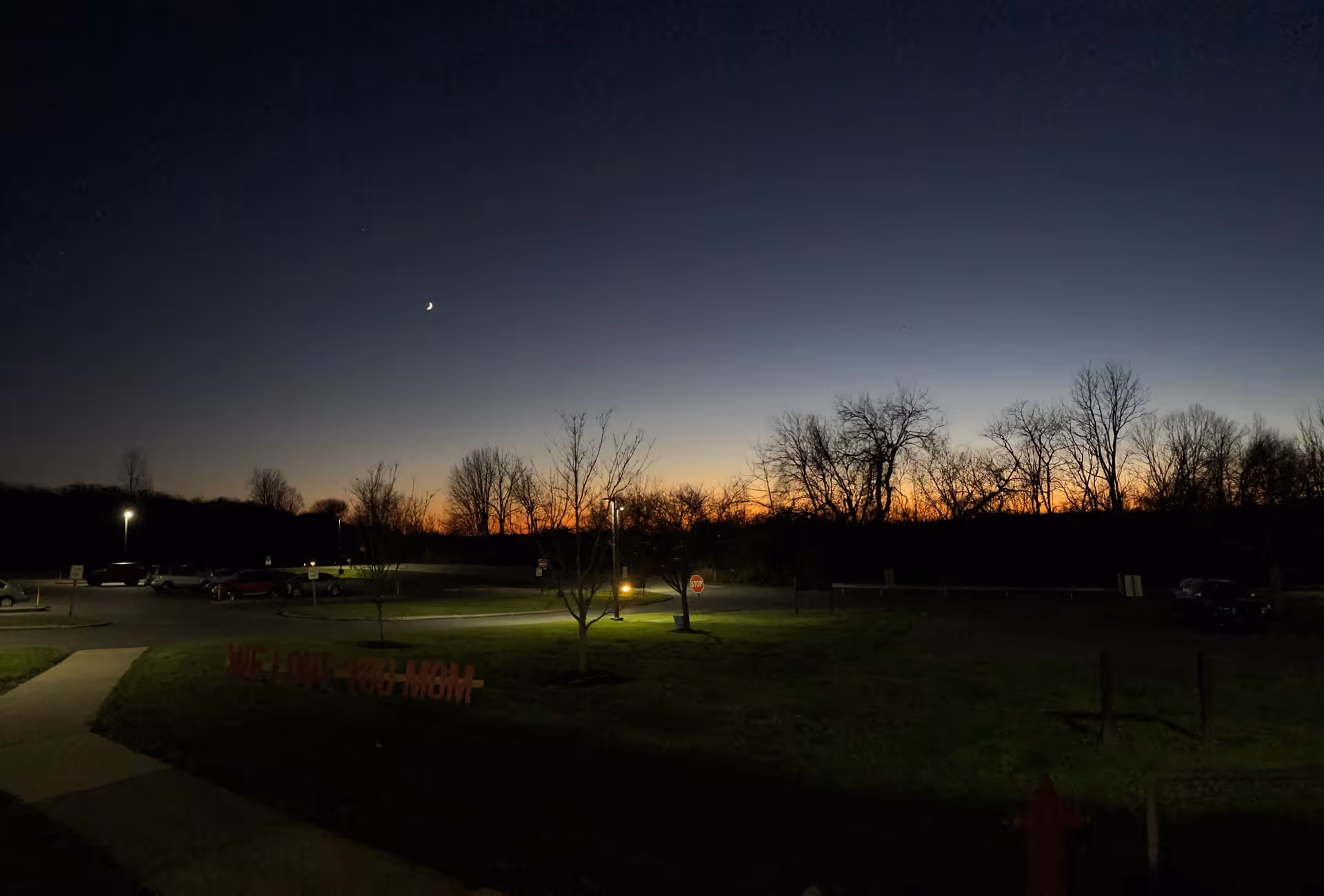 Parking lot and grassy lawn at dusk with a crescent moon, silhouetted trees, a lit walkway and a few parked cars.