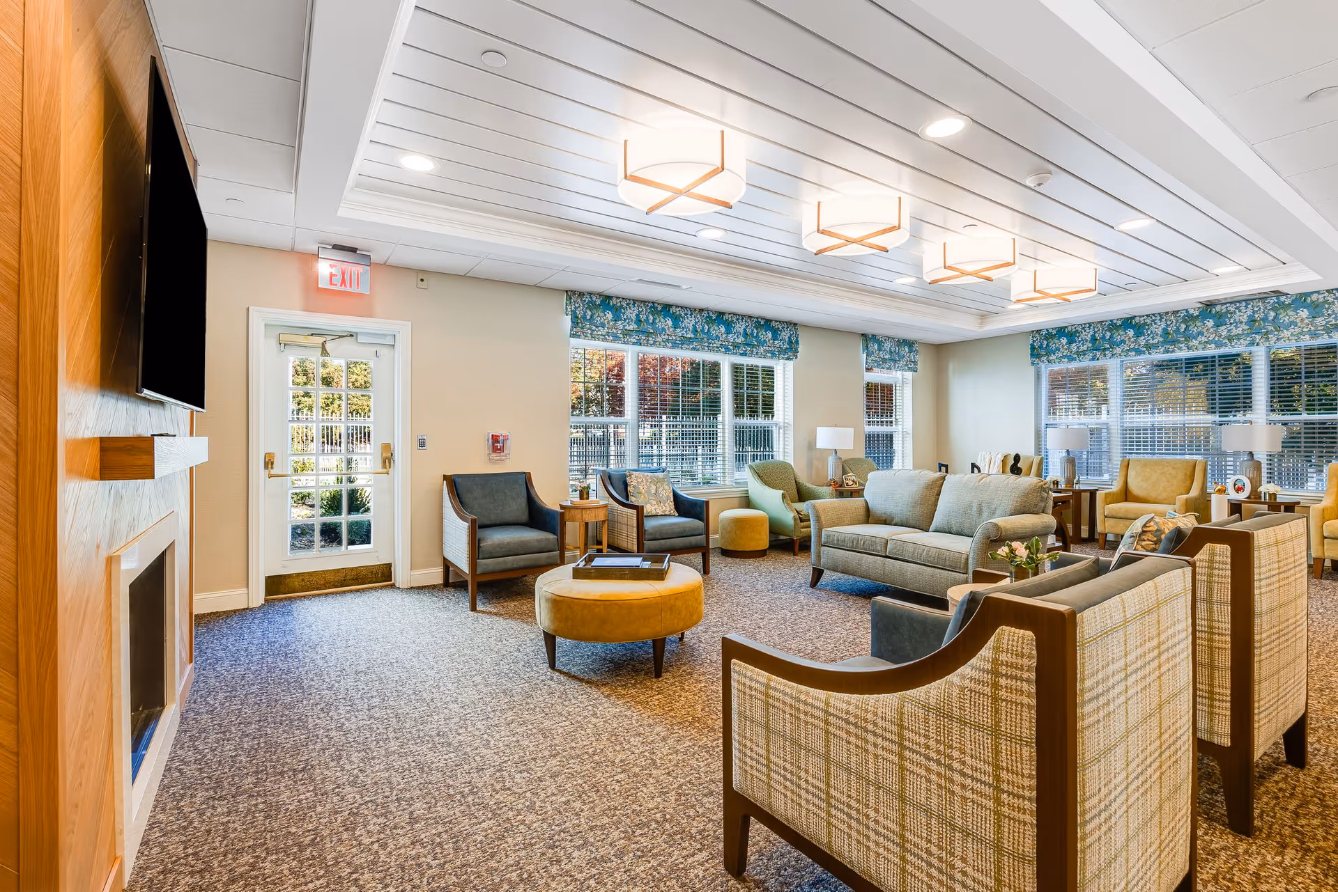 Spacious, well-lit senior living common room with sofas, armchairs, ottomans, and large windows.