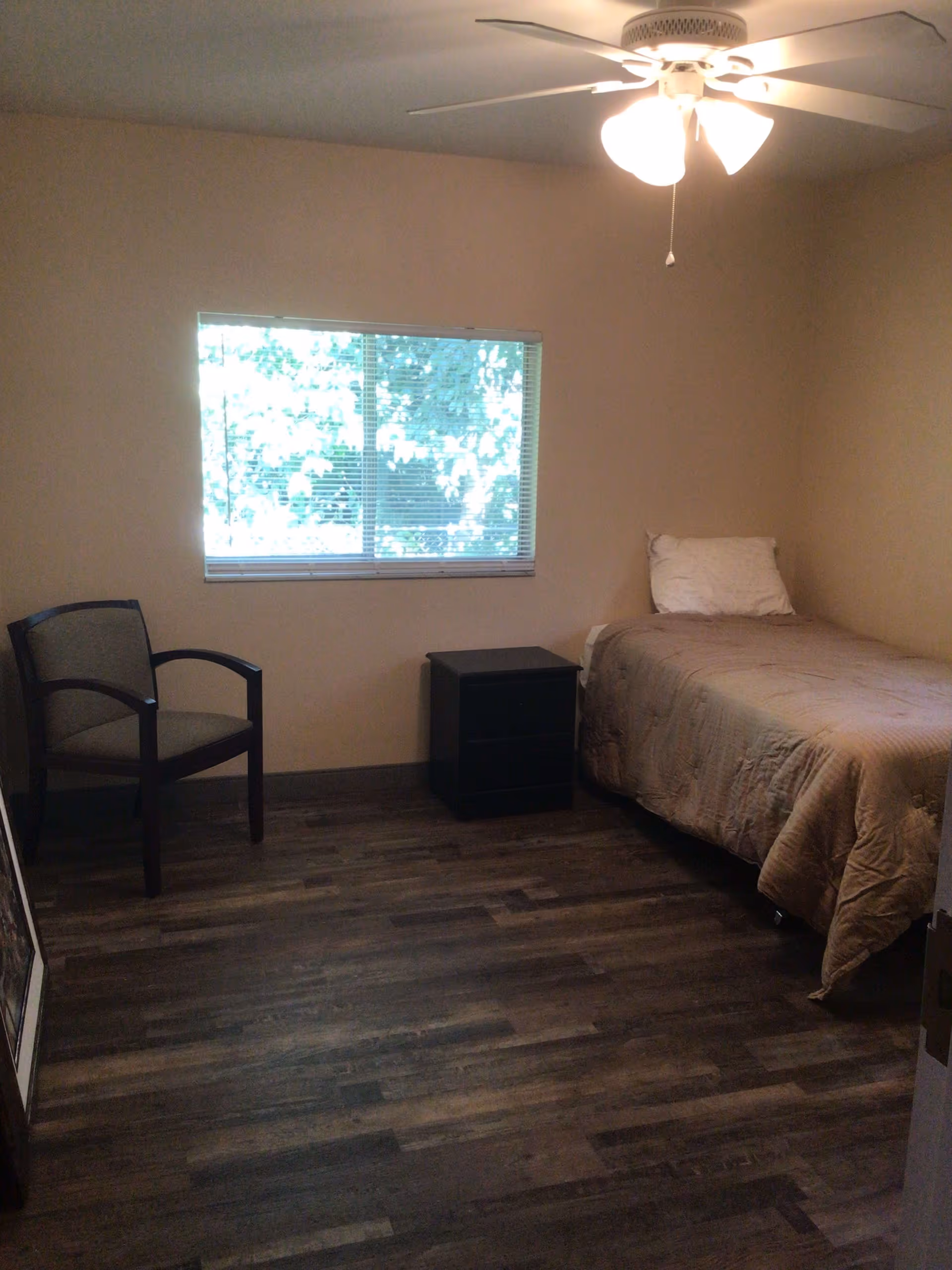 A simple bedroom with a single bed covered with a beige quilt and a white pillow. Next to the bed is a small black nightstand. A wooden chair with a cushioned seat and backrest is placed near a window with blinds. The room has wooden flooring and a ceiling fan with lights.