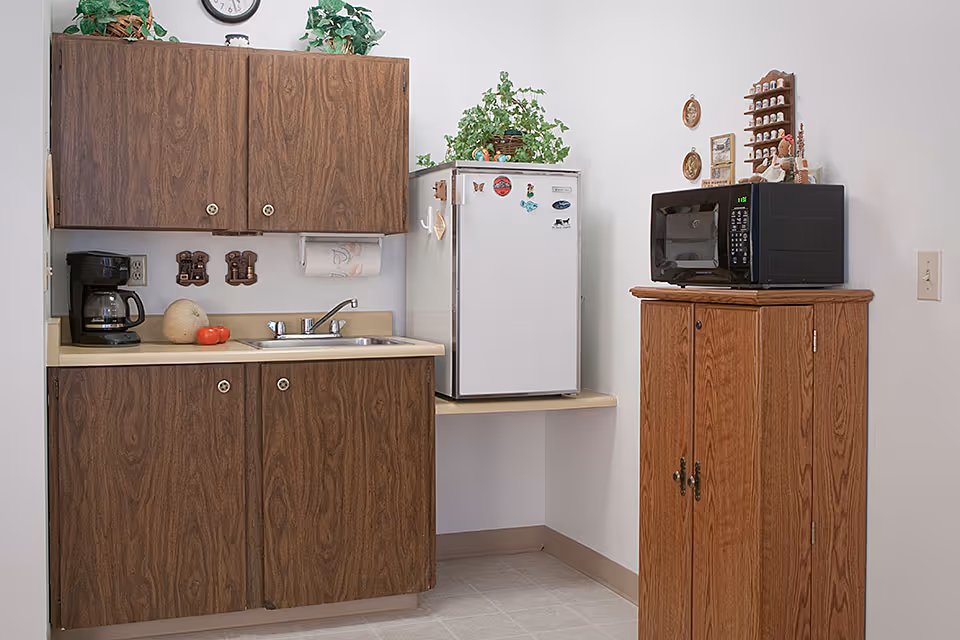 Small kitchenette area with wooden cabinets, a sink, a coffee maker, a white mini refrigerator with magnets, a microwave on a wooden cabinet, and some decorative plants and items on top of the cabinets.