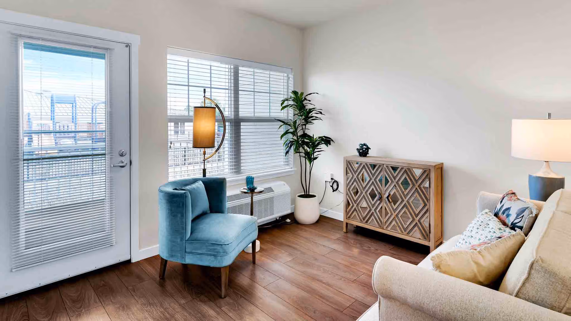 A bright living room with a beige sofa adorned with colorful pillows on the right, a wooden cabinet with a geometric pattern against the wall, a tall green potted plant, a blue upholstered chair next to a floor lamp with a yellow shade, and a door with window blinds leading outside.
