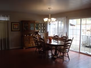 Dining room with a wooden dining table and six chairs. A chandelier hangs above the table. There is a wooden cabinet with glass doors against the wall, and framed pictures are displayed on the walls. Large sliding glass doors let in natural light from outside.