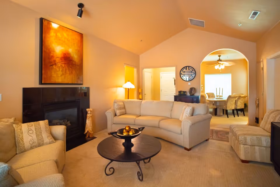A cozy living room with beige walls and carpet, featuring a beige sectional sofa, an armchair, and an ottoman. A round black coffee table with decorative items sits in the center. There is a black fireplace with an abstract orange and yellow painting above it. A floor lamp is lit next to the sofa. An arched doorway leads to a dining area with a table and chairs, illuminated by a ceiling fan with lights.