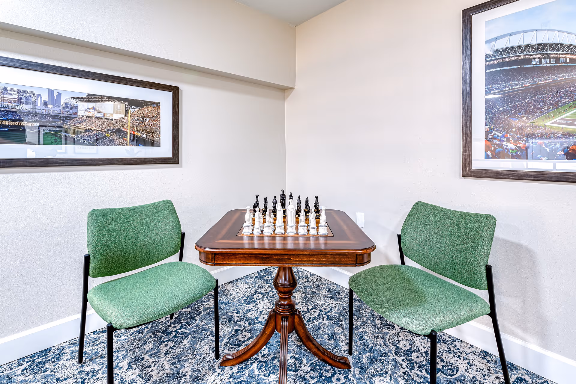 A small wooden table with a chessboard and chess pieces set up in the middle, flanked by two green upholstered chairs. The room has light-colored walls with two framed pictures of stadiums hanging on them, and a patterned blue and white carpet on the floor.