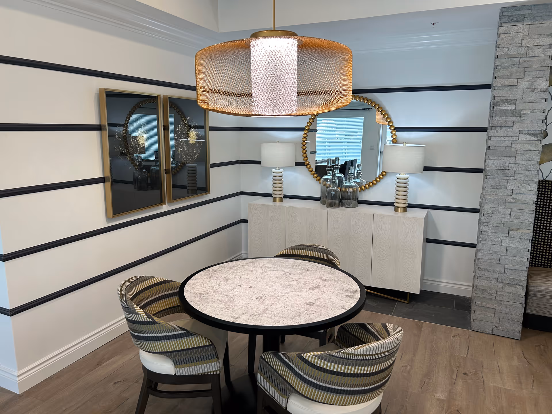 A modern dining area with a round marble table surrounded by four striped upholstered chairs. The walls have horizontal black stripes on a white background. A large round mirror with a gold beaded frame hangs above a white sideboard with two white and gold lamps and decorative glass bottles. A large, textured pendant light hangs above the table. The floor is a combination of wood and dark tiles, and a stone column is partially visible on the right.