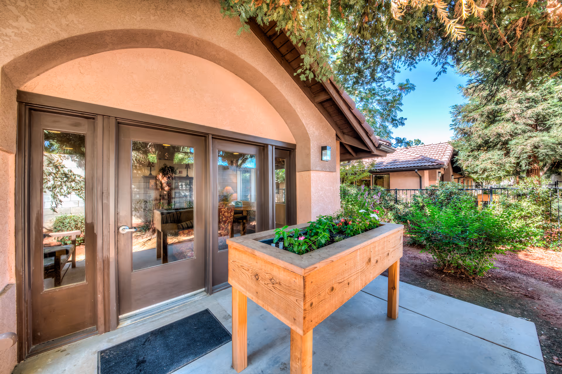 Outdoor patio area at Summerfield Memory Care Of Fresno featuring a raised wooden planter box with green plants and flowers, a concrete walkway, and a building with large glass doors and an arched entryway. Trees and shrubs surround the area under a clear blue sky.