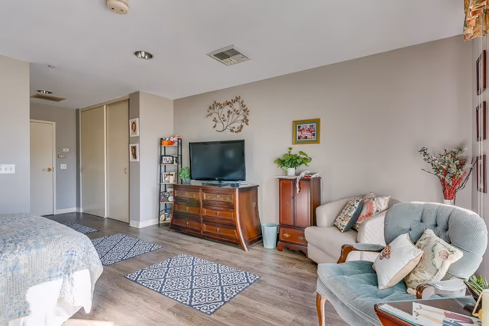 A cozy living space featuring a wooden dresser with a flat-screen TV on top, a small shelf with framed photos and decorative items, a beige loveseat with patterned pillows, a light blue upholstered armchair with cushions, and a bed partially visible on the left. The room has light gray walls, wood flooring, three patterned rugs, and a window with floral curtains letting in natural light.