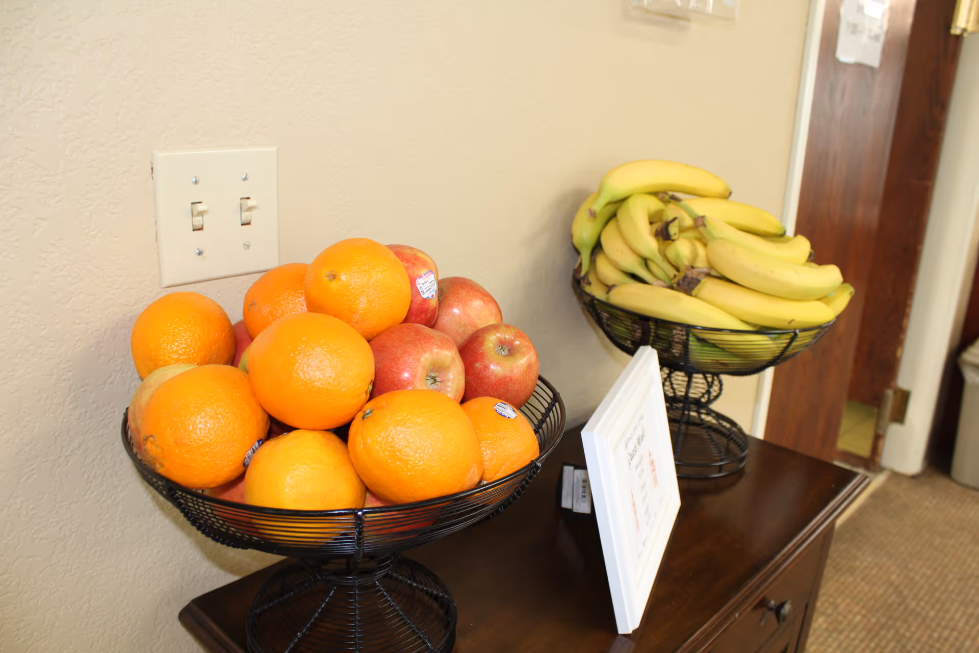 Two wire fruit bowls filled with oranges, apples, and bananas sit on a wooden table against a beige wall.