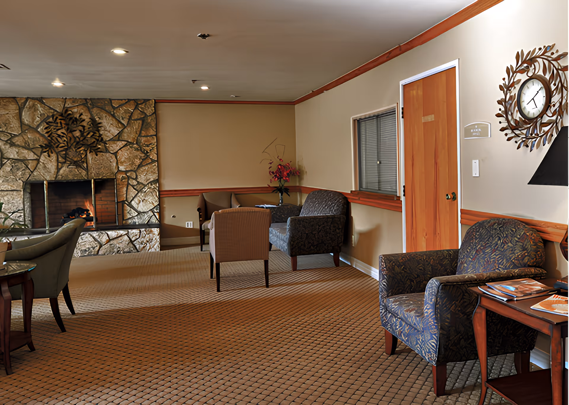A cozy lounge with upholstered chairs, side tables, and a stone fireplace in a senior living facility.