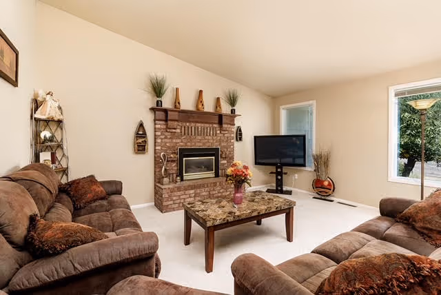 A cozy living room with brown cushioned sofas arranged around a rectangular coffee table with a floral centerpiece. The room features a brick fireplace with decorative items on the mantel, a flat-screen TV on a stand in the corner, a tall floor lamp near a window, and light-colored walls and carpet.