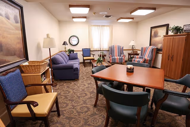 A cozy living room area with a patterned carpet, a wooden table surrounded by four green chairs, a blue couch, two striped armchairs, a wooden cabinet, and several lamps. The walls are decorated with framed paintings and a clock, and a window with light curtains lets in natural light.