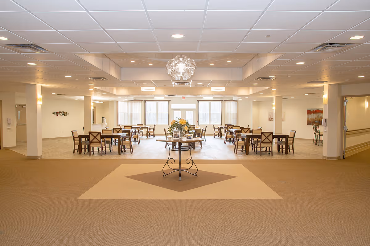 Spacious and well-lit dining area with multiple wooden tables and chairs arranged neatly. A round table with a flower arrangement is centered in the foreground. Large windows at the back allow natural light to fill the room, and the ceiling has recessed lighting and a decorative light fixture above the central table.