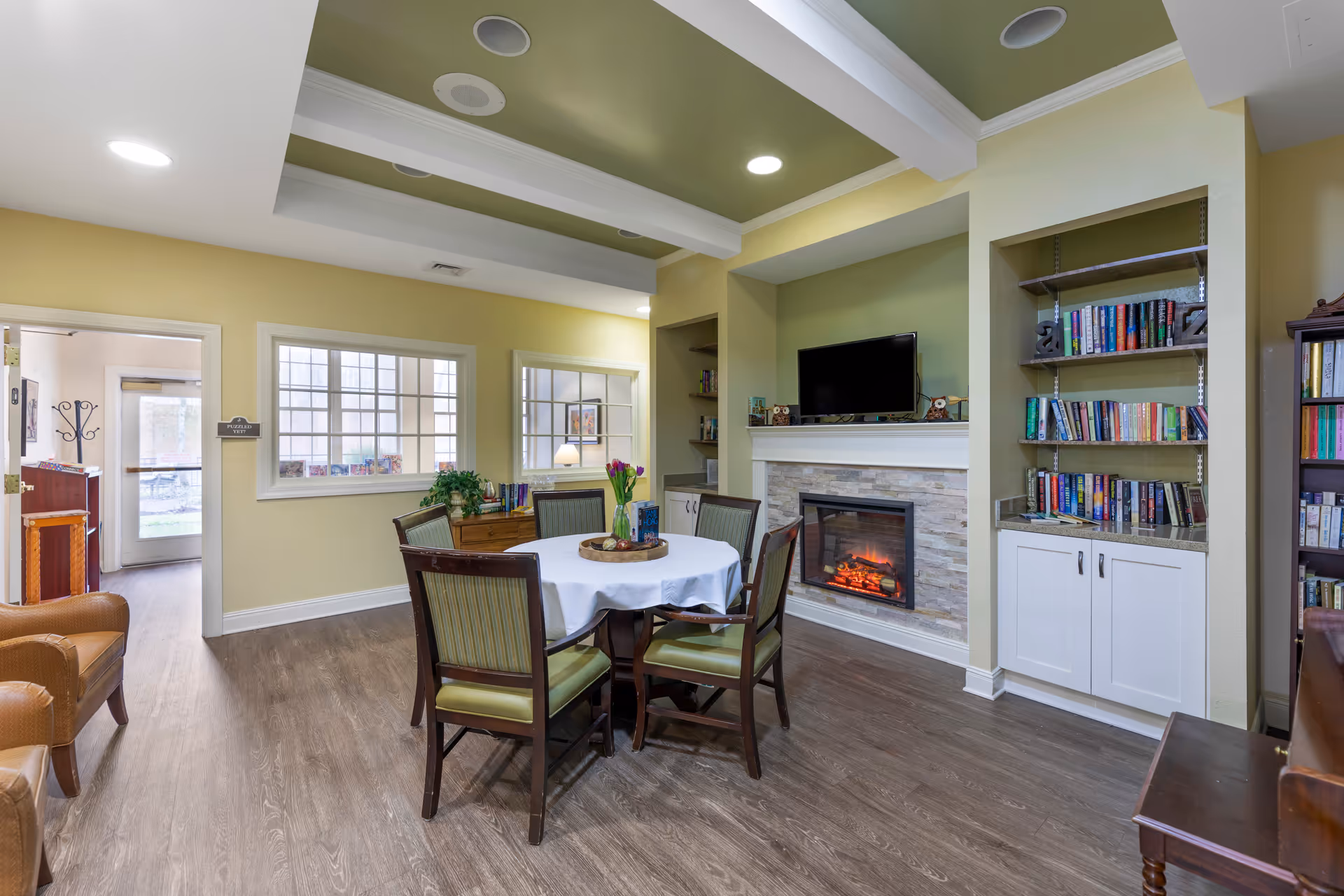 A cozy common area with a round table covered with a white tablecloth and surrounded by six chairs. There is a fireplace with a TV mounted above it, built-in bookshelves filled with books, and a wooden floor. The walls are painted light yellow and the ceiling has white beams with recessed lighting. A doorway leads to another room with a glass door and windows.