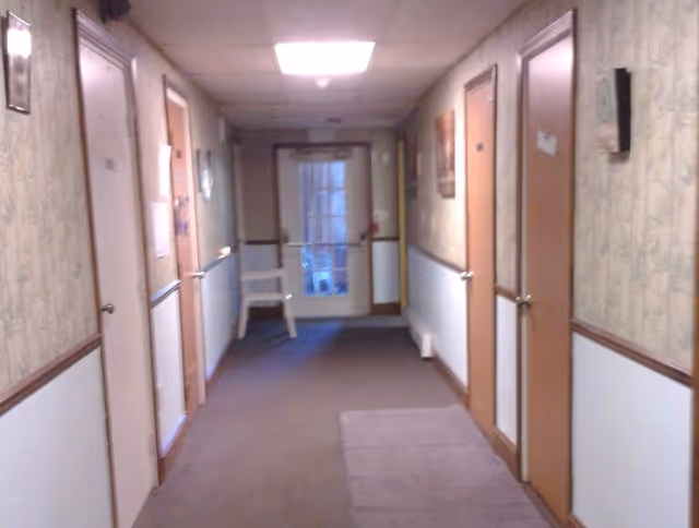 A carpeted hallway in a senior living facility with several closed wooden doors on both sides. The walls have a two-tone design with wallpaper on the upper half and white paneling on the lower half. A white chair is placed near a glass door at the end of the hallway, and there are framed pictures hanging on the walls.