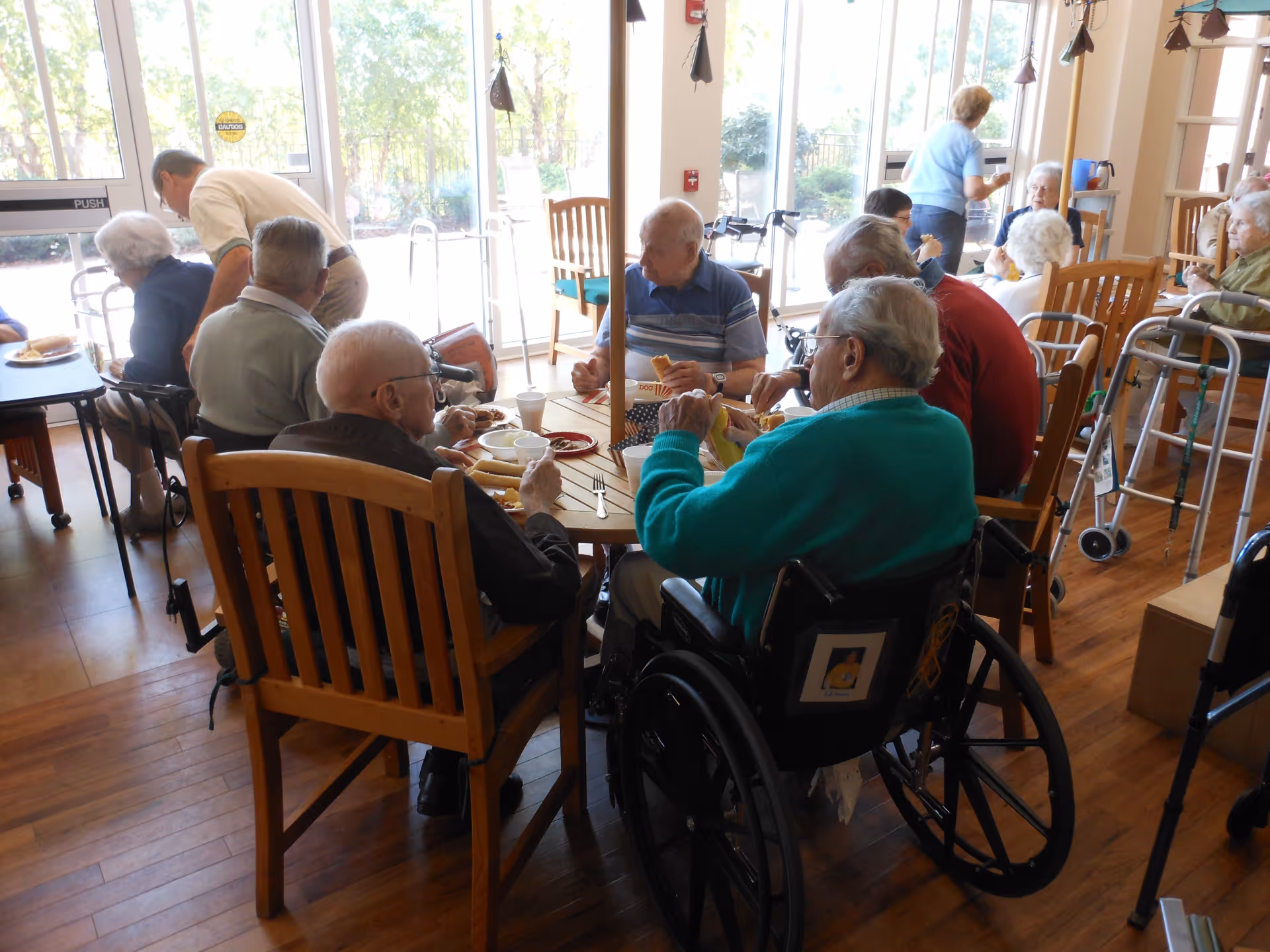 A group of elderly people sitting around tables in a dining area, eating and socializing. Some individuals are in wheelchairs, and others use walkers. The room has large windows letting in natural light and wooden flooring.