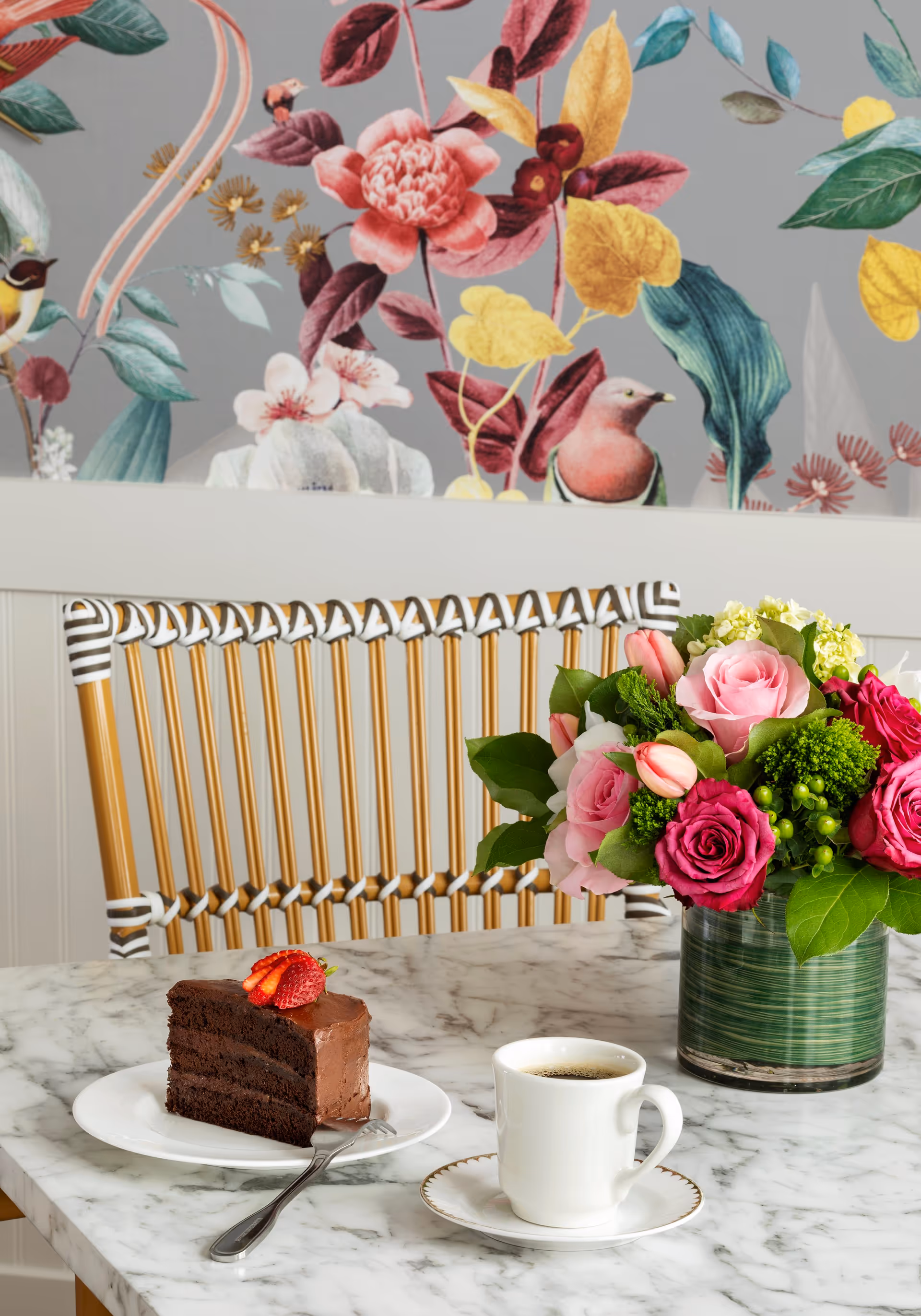 A marble table with a slice of chocolate cake topped with sliced strawberries on a white plate with a fork, a white cup of coffee on a saucer, and a green vase filled with pink and red roses and other flowers. Behind the table is a chair with a woven backrest and a wall decorated with floral and bird wallpaper.