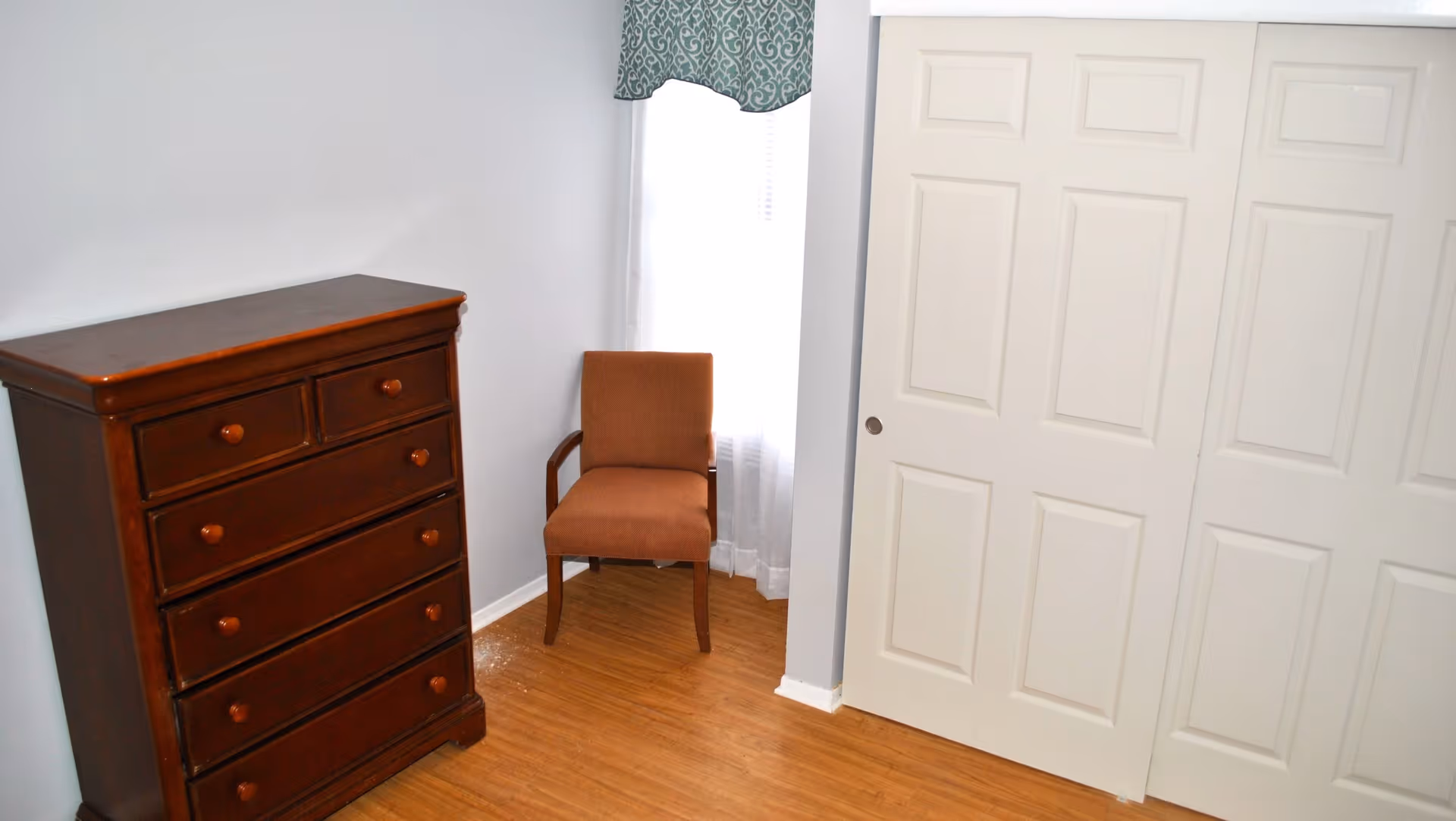 A simple room with a wooden chest of drawers on the left, a brown upholstered chair in front of a window with white curtains and a green patterned valance, and white sliding closet doors on the right. The floor is wooden and the walls are painted light gray.