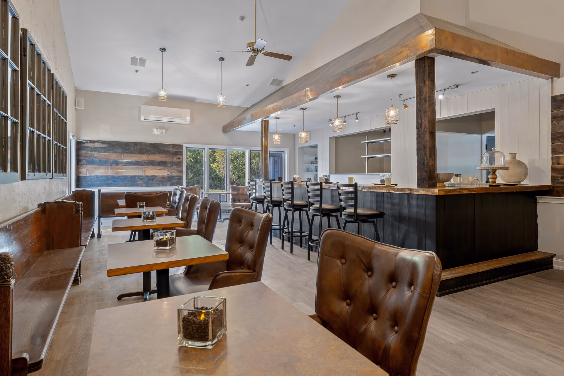 Interior view of a dining area in a senior living facility with wooden tables, brown leather chairs, a long wooden bench along the wall, and a bar counter with high stools. The room has large windows letting in natural light, pendant lights hanging from the ceiling, and a ceiling fan.