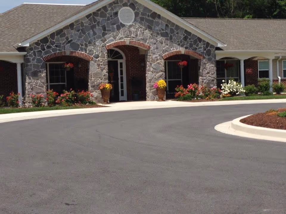 Front exterior view of a building with stone and brick facade, featuring three arched openings with hanging flower pots and large flower pots with colorful flowers near the entrance. The building is surrounded by a paved driveway and landscaped flower beds.