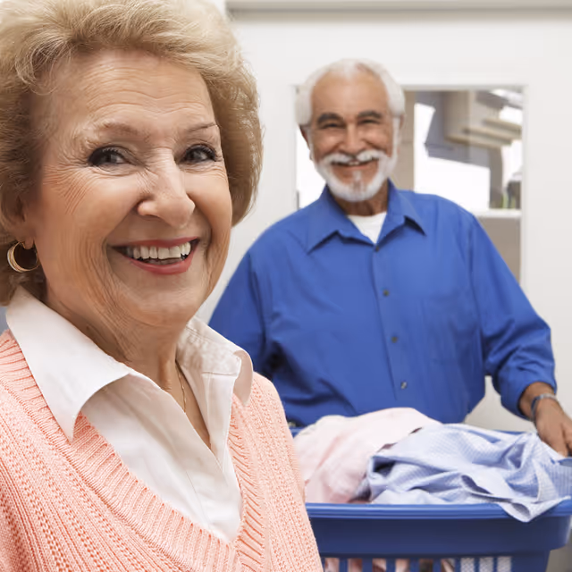 Smiling elderly woman in a pink sweater and white collared shirt stands in the foreground, while an elderly man in a blue shirt stands behind her holding a laundry basket filled with clothes in a bright indoor setting.
