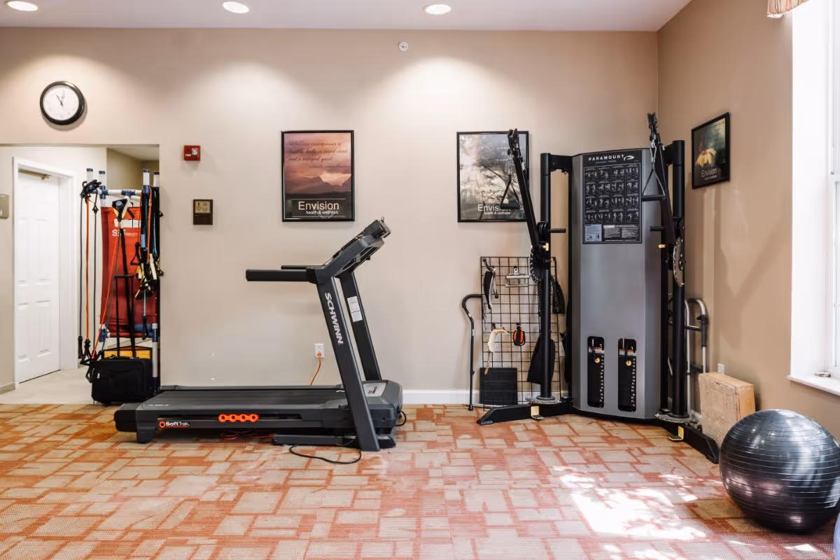 A fitness room with a treadmill, multi-exercise machine, exercise ball, and various fitness accessories against beige walls with framed motivational posters.