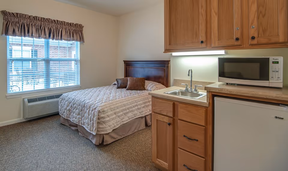 A cozy senior living room featuring a neatly made bed with two brown pillows, a window with blinds and a brown valance, a small kitchenette with a sink, wooden cabinets, a microwave, and a mini refrigerator.