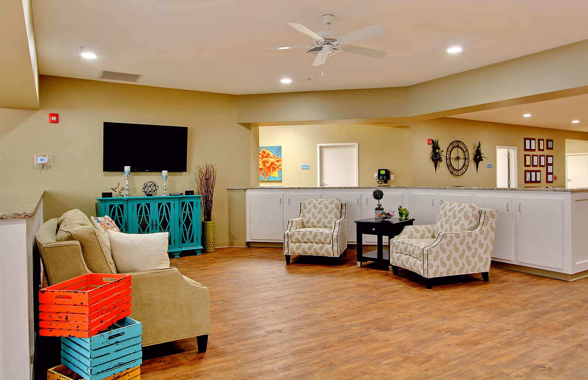 A spacious living room area in a senior care facility with wooden flooring, beige walls, and recessed lighting. The room features a beige sofa with pillows, two patterned armchairs around a small black table, a turquoise cabinet with decorative items, a wall-mounted flat-screen TV, and colorful wooden crates stacked in the corner. There is a ceiling fan above and white cabinetry along the back wall with decorative wall art and a clock.