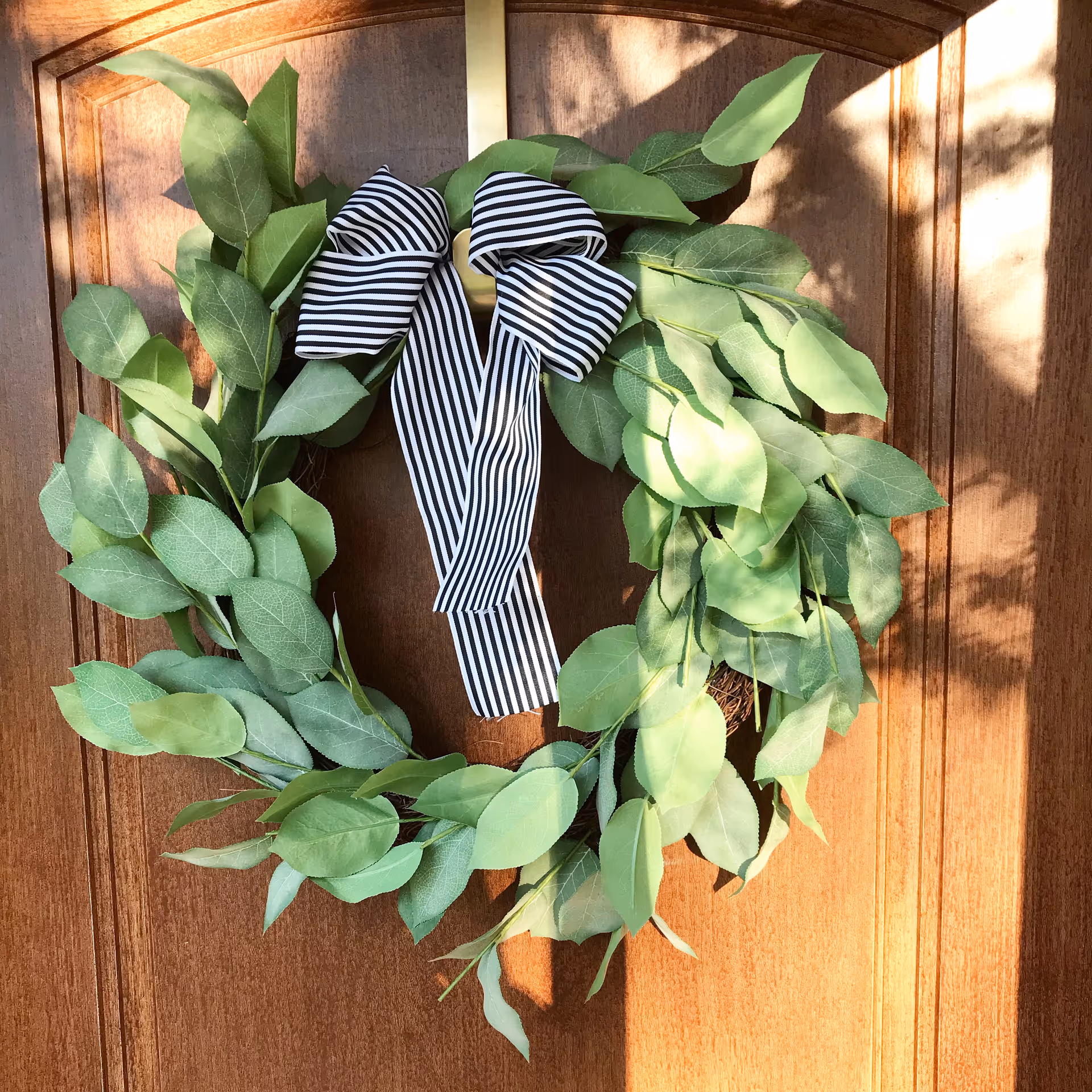 Green leafy wreath with a black-and-white striped bow hanging on a wooden front door.